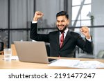 Small photo of Excited indian or arabian businessman in suit, sits in a modern office, looking at laptop, happy by good news, rejoice by win, raising hand in yes gesture, celebrating project success, big profit