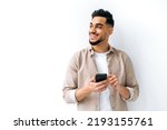 Small photo of Confident positive handsome indian or arabian young man, holding smartphone in hand, chatting online, browsing internet, looking happily to the side, standing on isolated white background, smiling