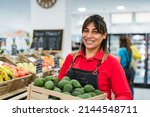 Small photo of Latin woman working in supermarket holding a box containing fresh avocados