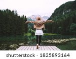 Young girl enjoying beauty of nature looking at mountain lake in Jezersko. Adventure travel in Slovenia, Europe. Woman stands with raised arms on wooden bridge on background with forest and Alps.