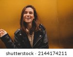 Simpering young brunette woman with a coy smile as she flirts over a gold studio background with copy space. Woman wear black leather jacket and beige shirt is laughing. 