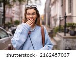 Astonished and shocked young woman in casual clothes covering her mouth with hand, looking at camera with amazed smiling facial expression walking on the street, urban. Human positive emotions concept