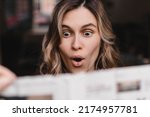 Shocked casual woman reading newspaper and look surprised while standing at home. Shocked curly girl look amazed, when read news in newspaper. Big eyes and open mouth.
