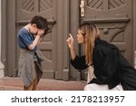 Small photo of Mother scolds her son on the street. Child cries, a woman shakes her finger because of the boy bad behavior, while walking to home. Rule of conduct. Woman sitting, boy cover his face and cry.