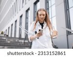 Small photo of Young confident business woman walking in hurry downstairs, holding mobile, glasses, coffee cup, texting on mobile phone on city street in front of modern office building. Serious busy woman.