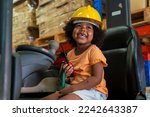 Small photo of three-year-old African-American girl in an engineer's helmet smiling happily drives a forklift as an engineer in a factory.