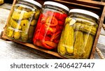 Small photo of Glass jars with pickled red bell peppers and pickled cucumbers (pickles) isolated in wooden crate. Jars with variety of pickled vegetables. Preserved food concept in a rustic composition.