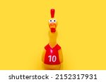Small photo of Rubber toy in the form of a crowing rooster on a yellow background in the center. Funny toy rooster has a surprised and dazed look with open beak and frightened eyes
