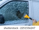 Small photo of A man in yellow winter jacket uses ice scrapers to thaw car windshield. Preparing car for driving after icy snowfall.