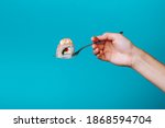 Hand holding fresh kunsan sushi roll with a fork, isolated on blue background. How to eat sushi without chopsticks