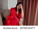 Small photo of Asian woman in a red satin nightgown and red long full length robe with headache, she is sitting on the gray sofa and touching her forehead. Thai woman dizzy at night.