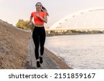 Young happy focused fitness girl in black yoga pants and orange short shirt jogs on riverbank during the day. Front view.