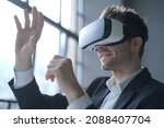 Small photo of European man office employee wears formal suit and VR headset glasses excited of experiencing virtual reality stretching his hands up trying to catch seemingly round object totally immersed in 3D game