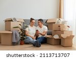 Small photo of Indoor shot of young couple in love moving in a new apartment, sitting on the floor near, planning to redecorate their new home, man holding smart phone, showing ideas to his wife.