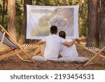 Small photo of Horizontal shot of young loving couple sitting in the forest with overhead projector, hugging each other and looking at projector screen, spending time in open air in beautiful nature.