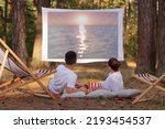 Small photo of Portrait of relaxed loving couple lying in the forest with overhead projector, watching romantic movie, enjoying sunset on screen and beautiful nature in the wood.