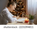 Indoor shot of young adult woman writing resolutions for New year in notebook at home, sitting by Christmas tree, wearing white shirt. Motivation for future.