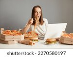 Indoor shot of busy hungry woman eating fast food while working on laptop, sitting at table against gray wall, working on laptop and eating pasta, keeps mouth open, looking at notebook display.