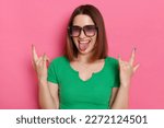 Indoor shot of excited playful crazy young girl with brown hair wearing green casual t shirt and sunglasses posing against pink wall, showing rock and roll gesture and tongue out