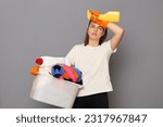 Housewife takes care of clothing. Domestic appliances aid in housework. Tired exhaused woman wearing whote t-shirt with laundry bowl and detergent isolated over gray background