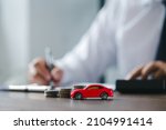 Small photo of Car on desk with stack coins and man signing purchase documents in background. Red modern car while hand completes the insurance policy or rental documents. Guy buying new car at dealership.