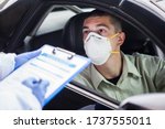 Small photo of Male driver wearing protective N95 face mask sitting by left drive wheel in UK drive-thru COVID-19 test centre,answering health check up questions,medical worker ticking off symptoms on clipboard form