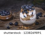 A healthy breakfast of yoghurt, homemade granola and blueberries in a glass. The glass is on a worn stone surface and next to the glass is a small bowl of blueberries. Some blueberries and granola are