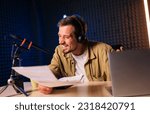 Small photo of Smiling and gesturing radio host with headphones reading news from paper into studio microphone at radio station with neon lights
