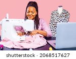 Small photo of african tailor with afro dreadlocks pigtails sews clothes on sewing machine at tailor office pink wall background