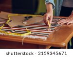 Small photo of Creative middle aged woman tailor making paper pattern with scissors for sewing clothes. Woman seamstress working from home and tailoring beautiful handmade dresses. Focus on tailors hand using chalk