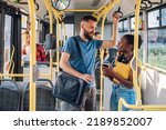 Cheerful diverse friends smiling and talking together and using a smartphone while riding a bus in the city and going to work. Standing in public transport, commuting by bus. Diverse romantic couple.