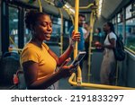 An attractive african american woman using a smartphone while riding a bus in the night. Young beautiful black woman using public transportation. Illuminated by the city light.