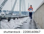 Small photo of Smiling female runner jogging on a bridge during winter. Woman in sportswear running on the bridge on a snow. Copy space. Urban city jogger. Training in a urban area.