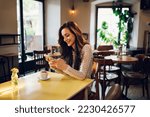Small photo of Young beautiful woman with natural makeup relaxing with cup of coffee ans using a smartphone while sitting at wooden table in a coffee shop. Smiling female typing on a mobile phone. Beauty concept.