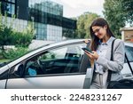 Portrait of a confident business woman wearing elegant suit and typing message on a smartphone while getting into the car. Businesswoman using mobile phone while standing next to open car door.