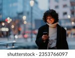 Wide shot of a joyful dark haired young woman standing on the street in a city center and looking down to her smart phone. Having a big smile.Blurred background of a street and city lights.Copy space.