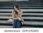 Small photo of Beautiful stylish woman sitting on a stairs outside and looking really sad. Fashionable female in trench coat with a bag put on a side looking away and looking melancholic. Copy space.