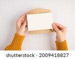 First person top view photo of female hands in yellow sweater holding open craft paper envelope and white card on isolated white background with blank space