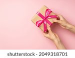 First person top view photo of female hands giving stylish craft paper giftbox with pink satin ribbon bow on isolated pastel pink background with empty space
