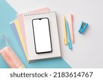 Small photo of School supplies concept. Top view photo of smartphone over notepads pens pencil-case and mini stapler on bicolor blue and white background with empty space