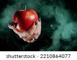 Small photo of Woman as witch offers red apple as symbol of temptation, poison. Fairy tale, white snow wizard concept. Spooky halloween, cosplay. Smoke, haze background.