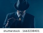 Mafioso with covered eyes with felt hat lighting cigar on dark blue background