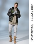 Small photo of full length of young stylish man in hat, hoodie and anorak standing on grey