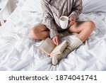cropped view of woman in sweater and knitted socks sitting in bed with mug of hot cocoa and book at morning
