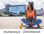 african american woman in sunglasses sitting with crossed legs on longboard near plastic cup with orange juice on asphalt