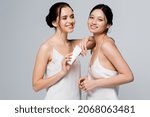 Happy multiethnic women in satin dresses holding cream in tube isolated on grey