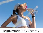 low angle view of sportive woman in baseball cap and sports bra drinking water against blue sky