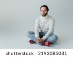 full length of bearded man in jeans and knitted sweater sitting in red socks with ornament on grey