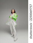 Small photo of full length of young asian woman in pants and green and white jacket looking at camera on grey background