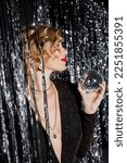 Small photo of side view of blonde woman with red lips holding small disco ball near tinsel curtain on grey